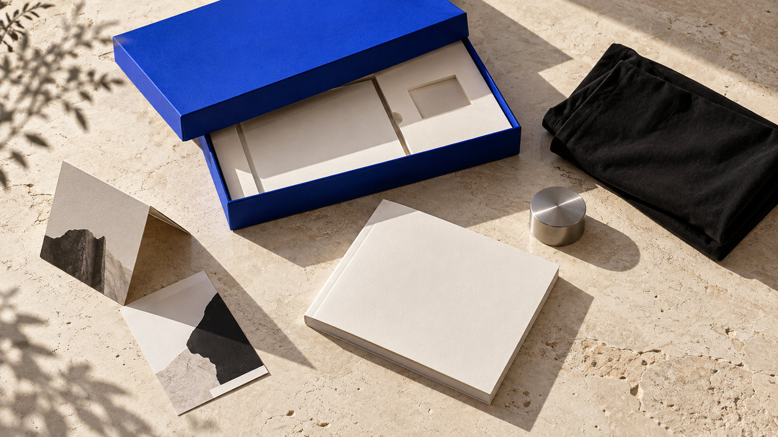 Blue presentation box, ivory booklet and printed cards with folded black fabric and a small steel object on limestone.